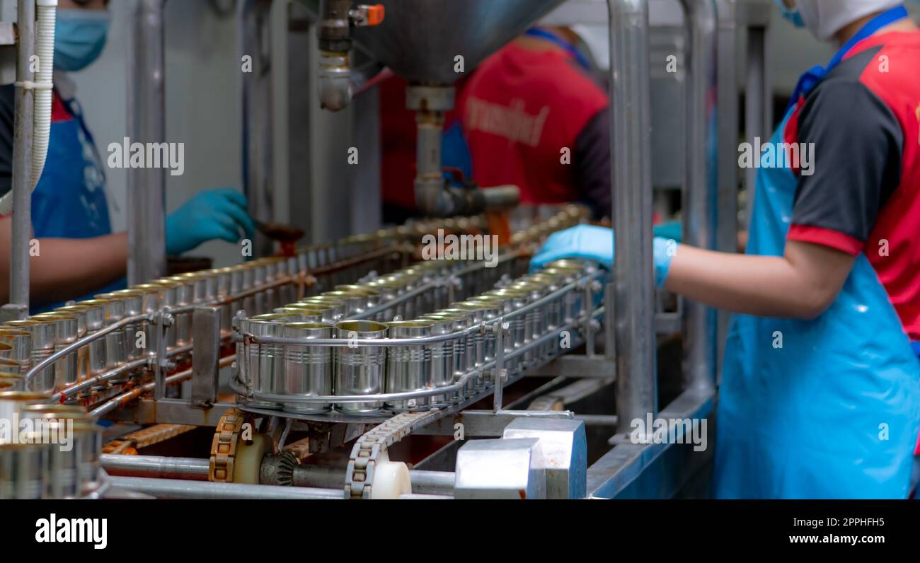 Canned fish factory. Food industry.  Sardines in red tomato sauce in tinned cans on conveyor belt at food factory. Blur workers working in food processing production line. Food manufacturing industry. Stock Photo