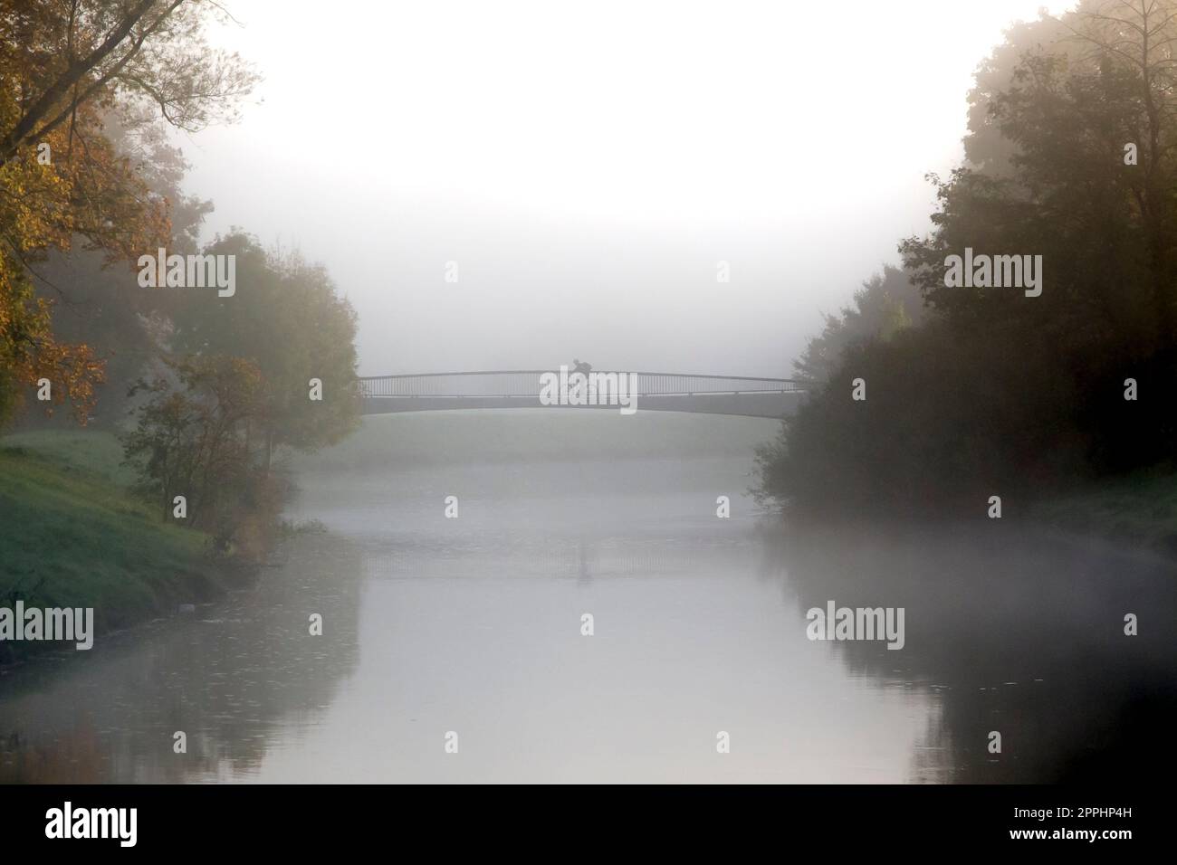 Lonely cyclist in morning or evening fog on bridge over small river Stock Photo