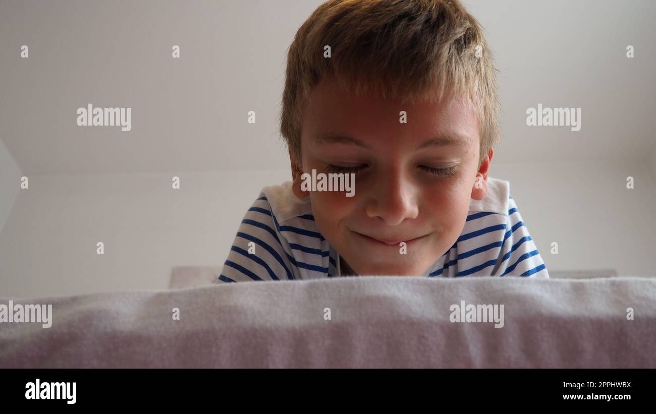 Cute little caucasian boy 10 years old lies at home on the bed in the bedroom and smiles slyly, squinting and closing his eyes. Expecting a surprise, a joke and pampering. Peekaboo. Play Blinders. Stock Photo