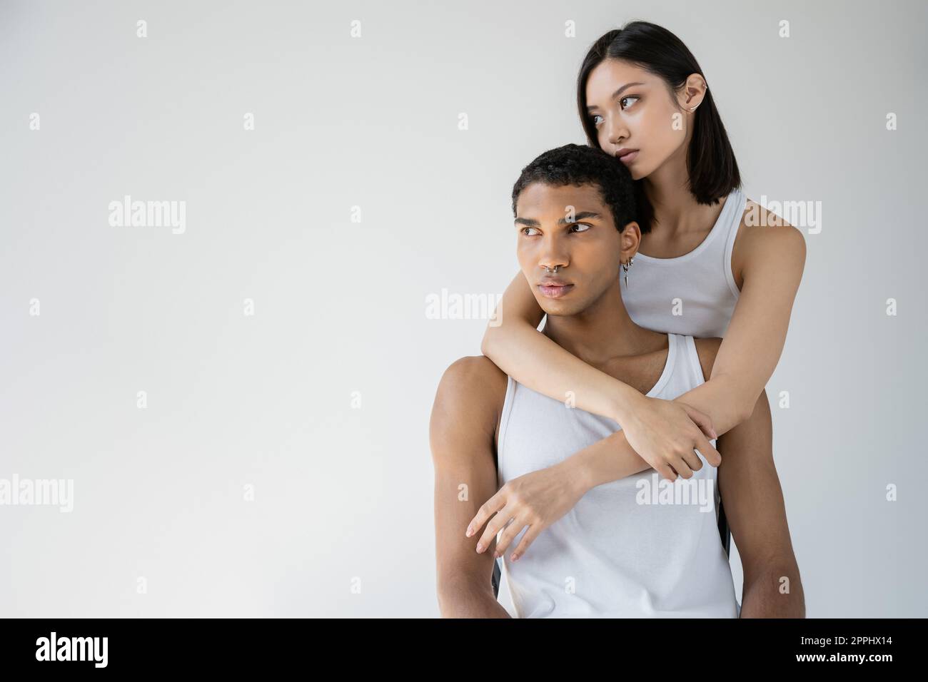 young asian woman looking away and hugging african american man in white tank top isolated on grey,stock image Stock Photo