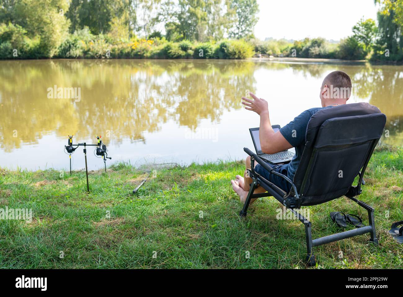 A man sits in a chair near the autumn lake and works on a laptop freelance camping. Robot out of the office, phoned with colleagues. Stock Photo