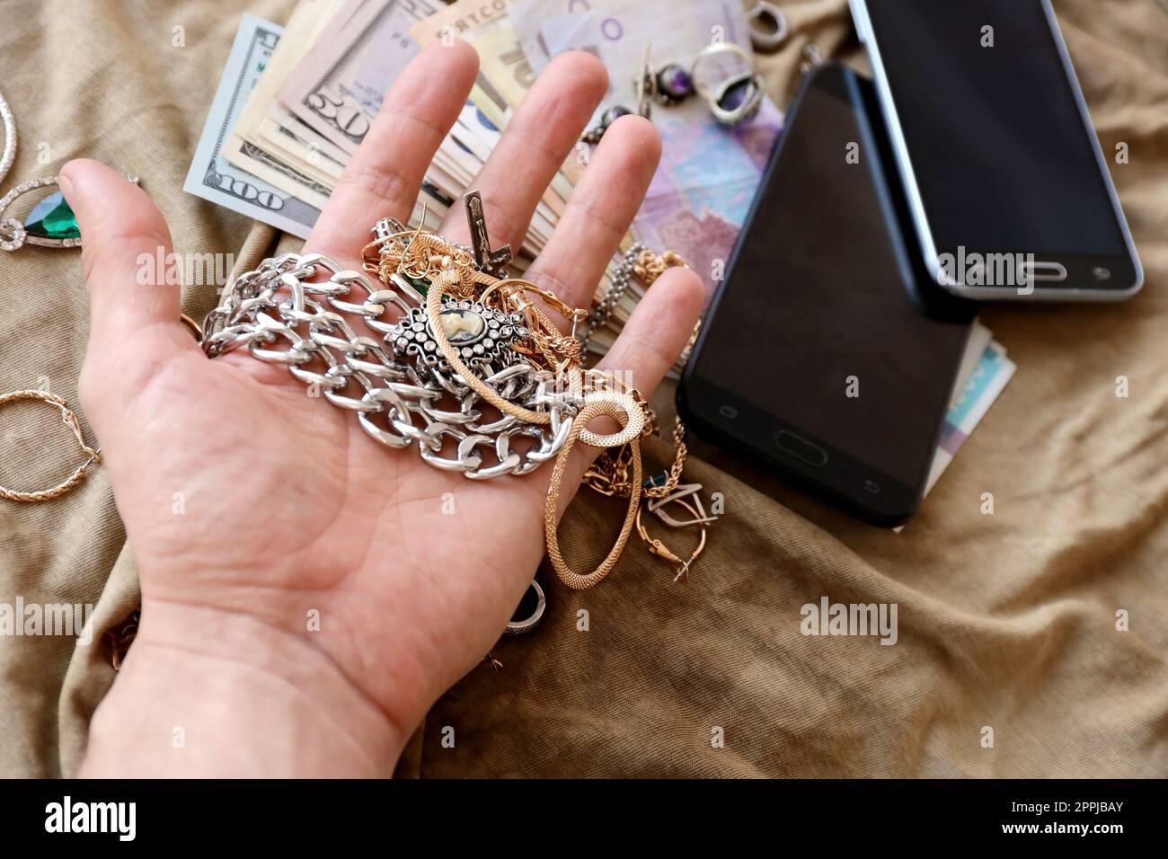 Marauders hand with bunch of stolen jewelry, money and smartphones on military uniform cloth fabric. Looting by Russian soldiers in the Ukrainian cities during Russian invasion on Ukraine Stock Photo