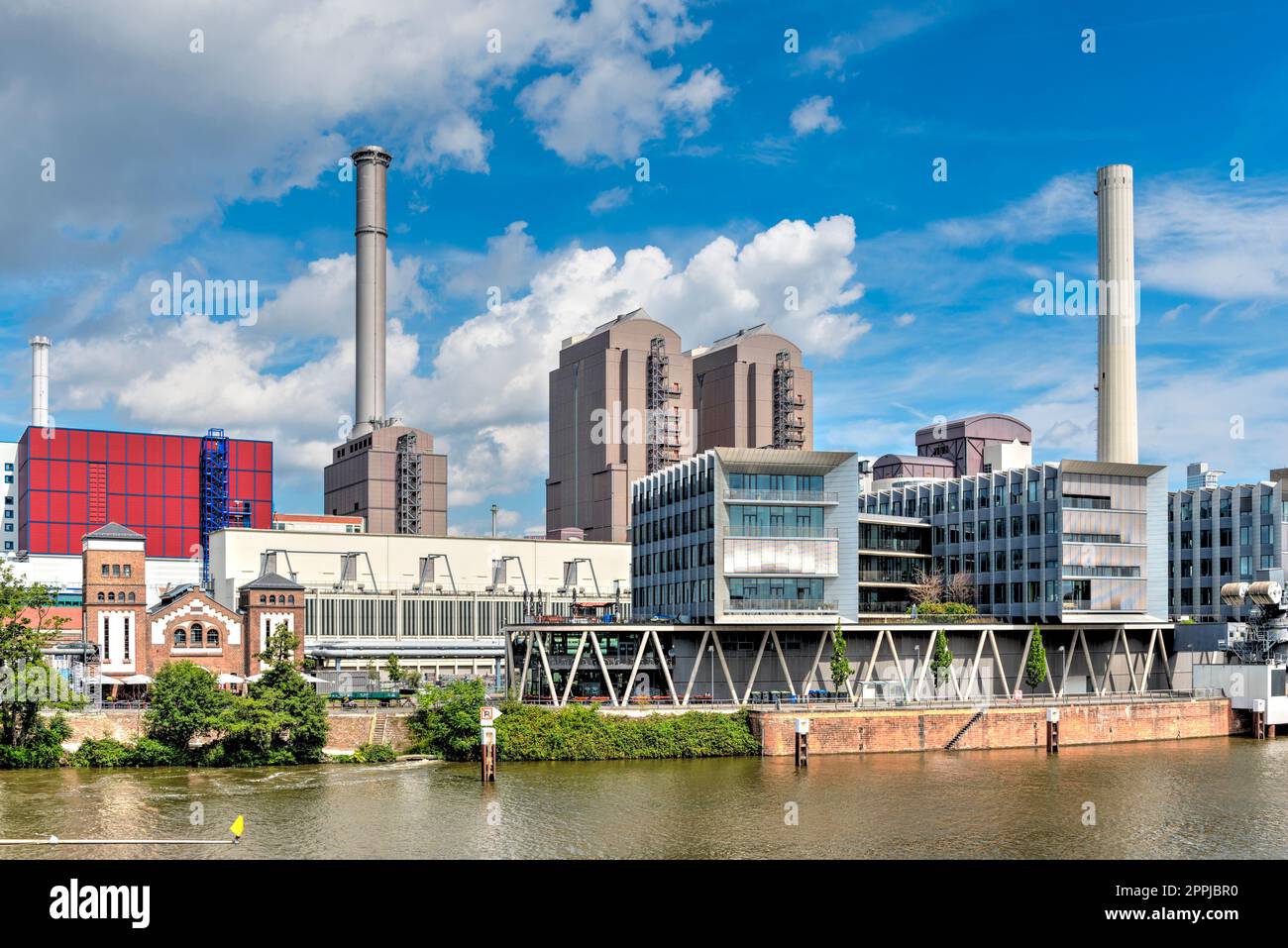 The bank of the Westhafen of Frankfurt am Main with combined heat and power plant, smokestacks, office buildings and industrial plants with the river Main in the foreground in summer weather with spring clouds Stock Photo