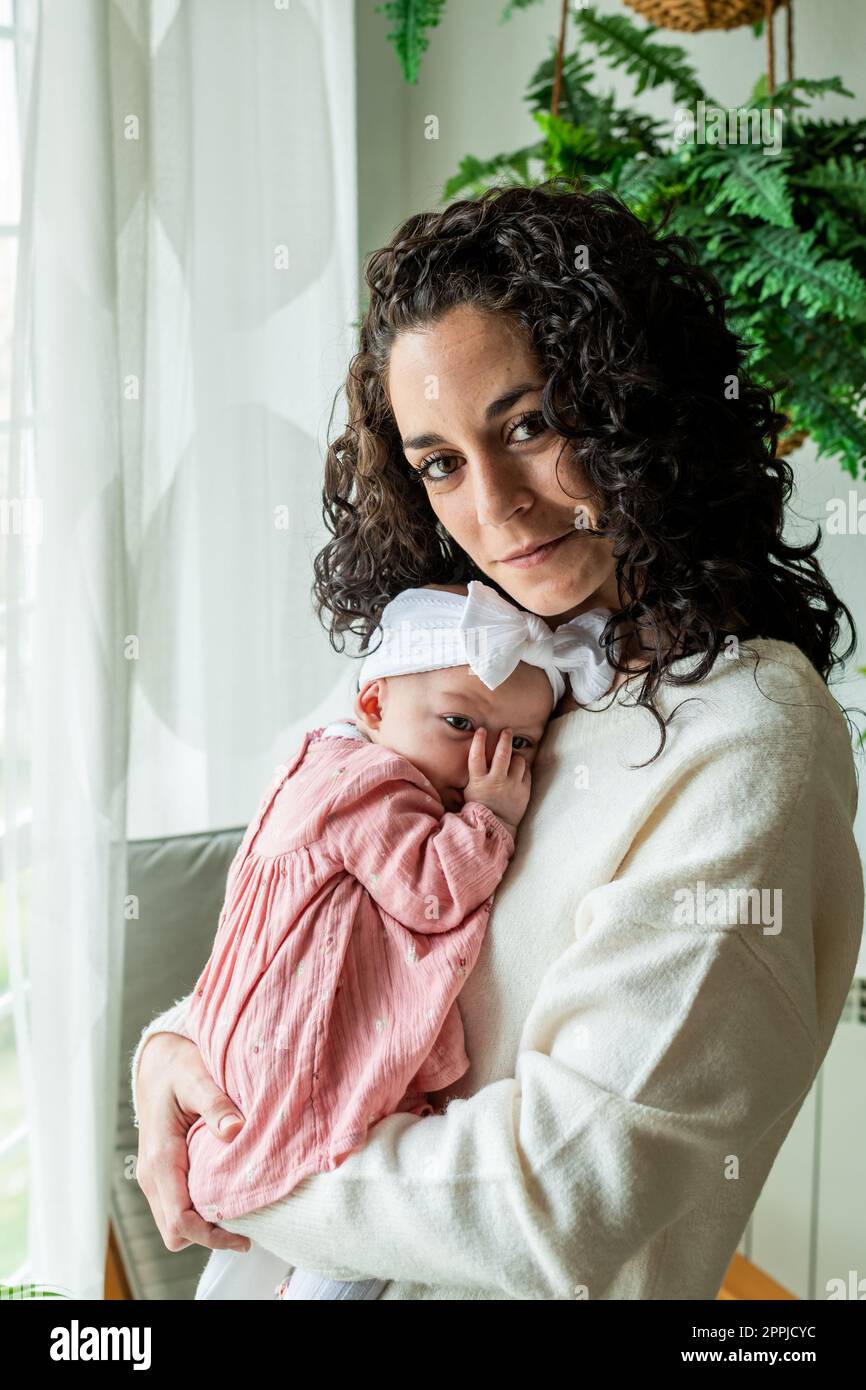 young mother with curly hair poses looking at camera while holding her newborn baby in her arms sucking his thumb. family and newborn care concept. Stock Photo