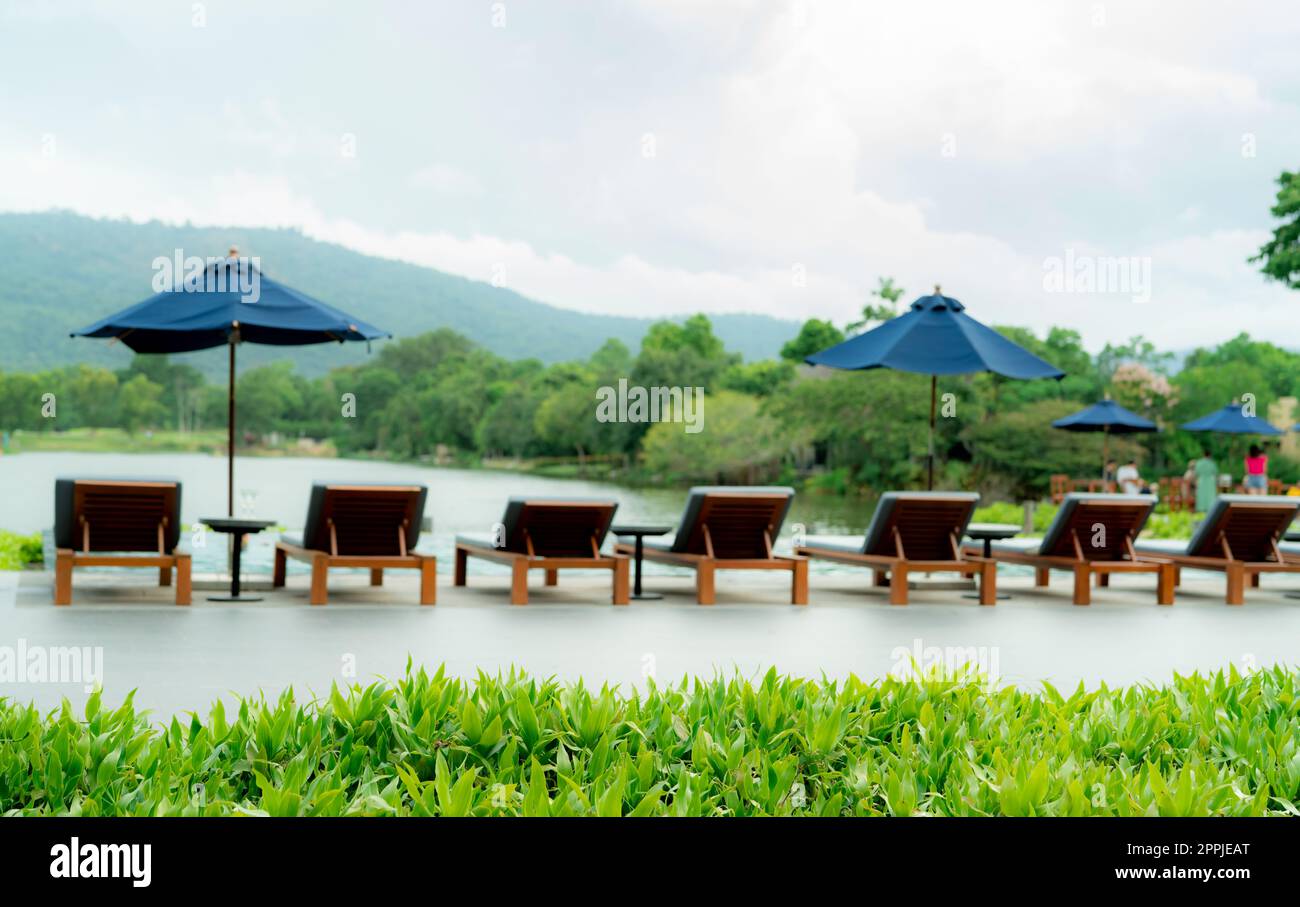Green grass on blur wooden beach chair and beach umbrella beside swimming pool at luxury hotel near the mountain. Summer vacation travel. Tropical holiday. Eco-friendly and sustainable hotel or resort Stock Photo
