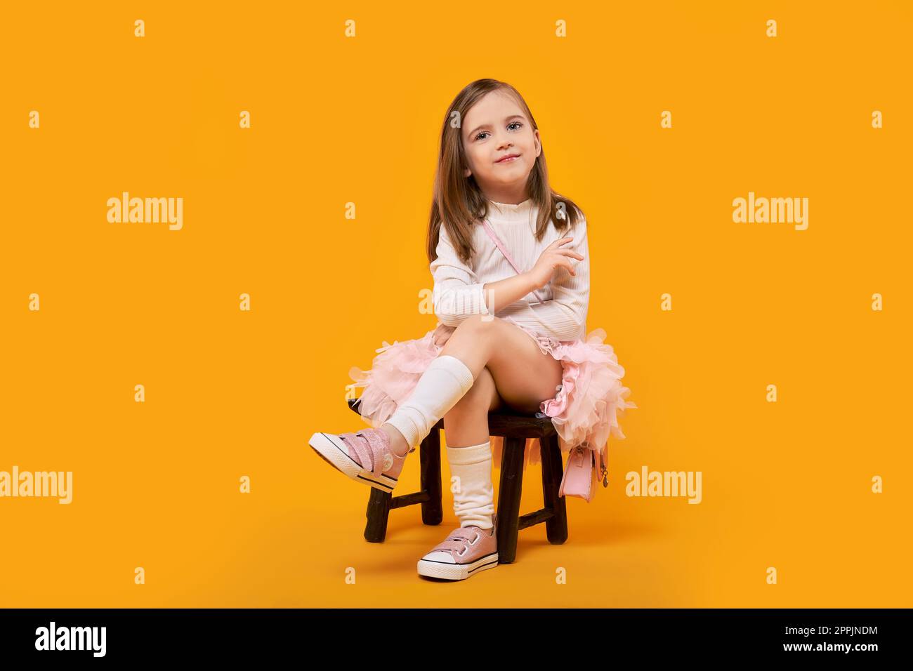 Young girl in tulle skirt and white pullover sitting on small wooden stool on bright yellow background Stock Photo