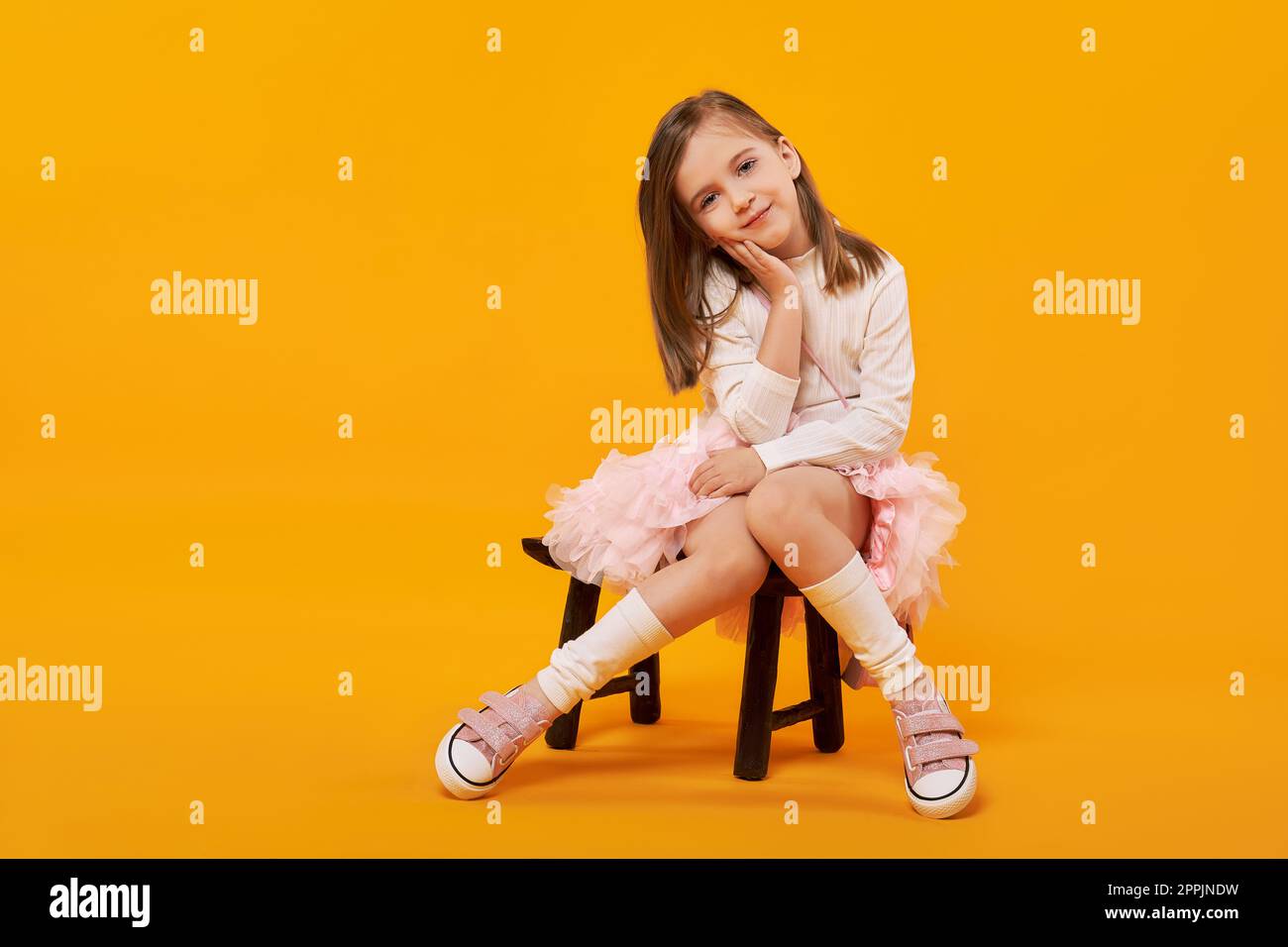 Lovely young girl sitting on small wooden chair in tulle skirt, white pullover and white gaiters Stock Photo