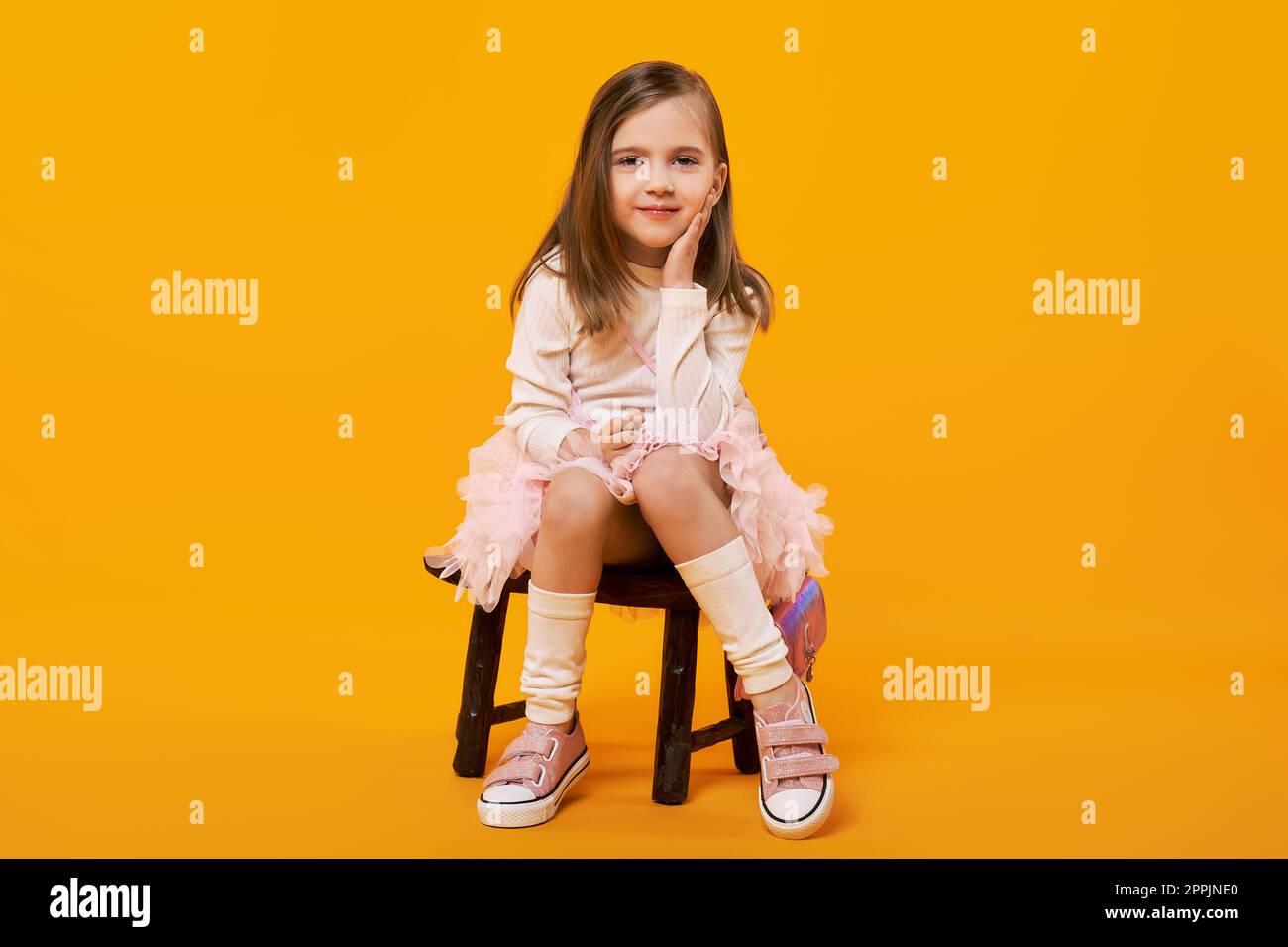 Young girl in tulle skirt and white pullover sitting on small wooden stool on bright yellow background Stock Photo