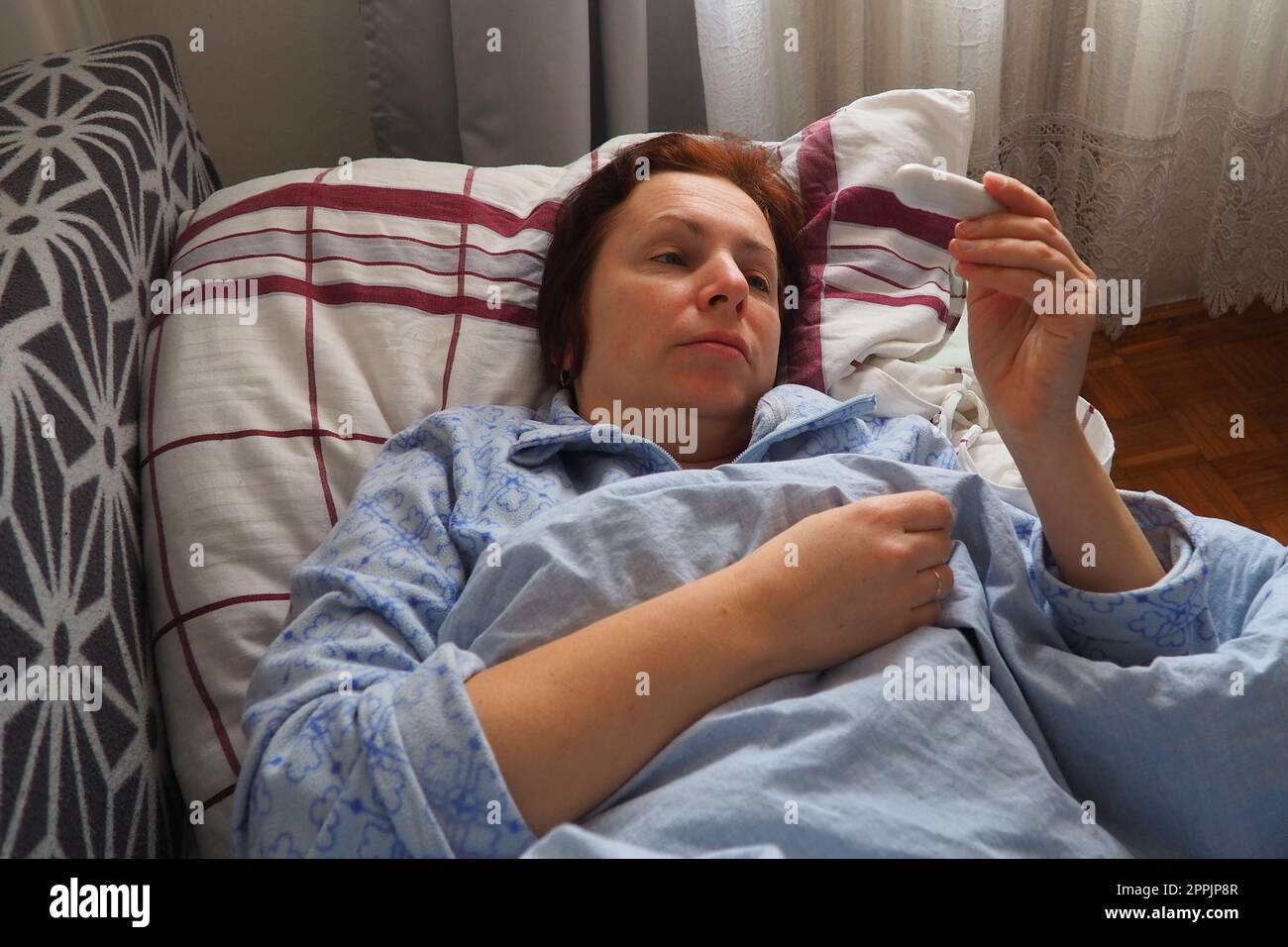 Sick woman in bed. Headache, runny nose and cold. Paleness of the face and faintness of the body. Feeling unwell. The patient lies on a pillow, holds a thermometer and measures the temperature. Stock Photo