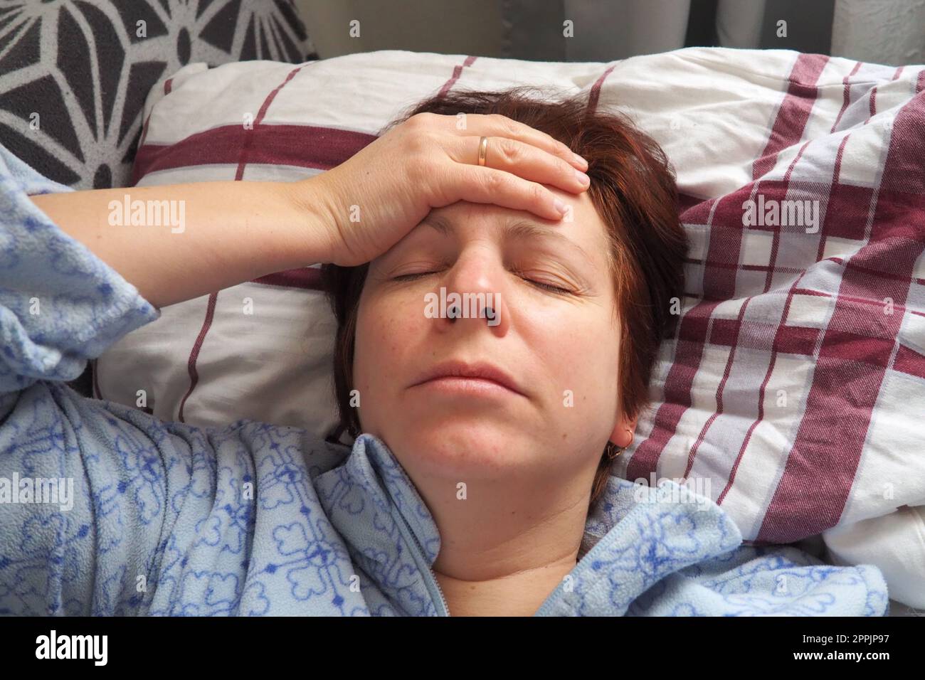 Sick woman in bed. Headache, runny nose and cold. Paleness of the face and faintness of the body. Feeling unwell. The patient holds his forehead with his hand and covers his eyes Stock Photo