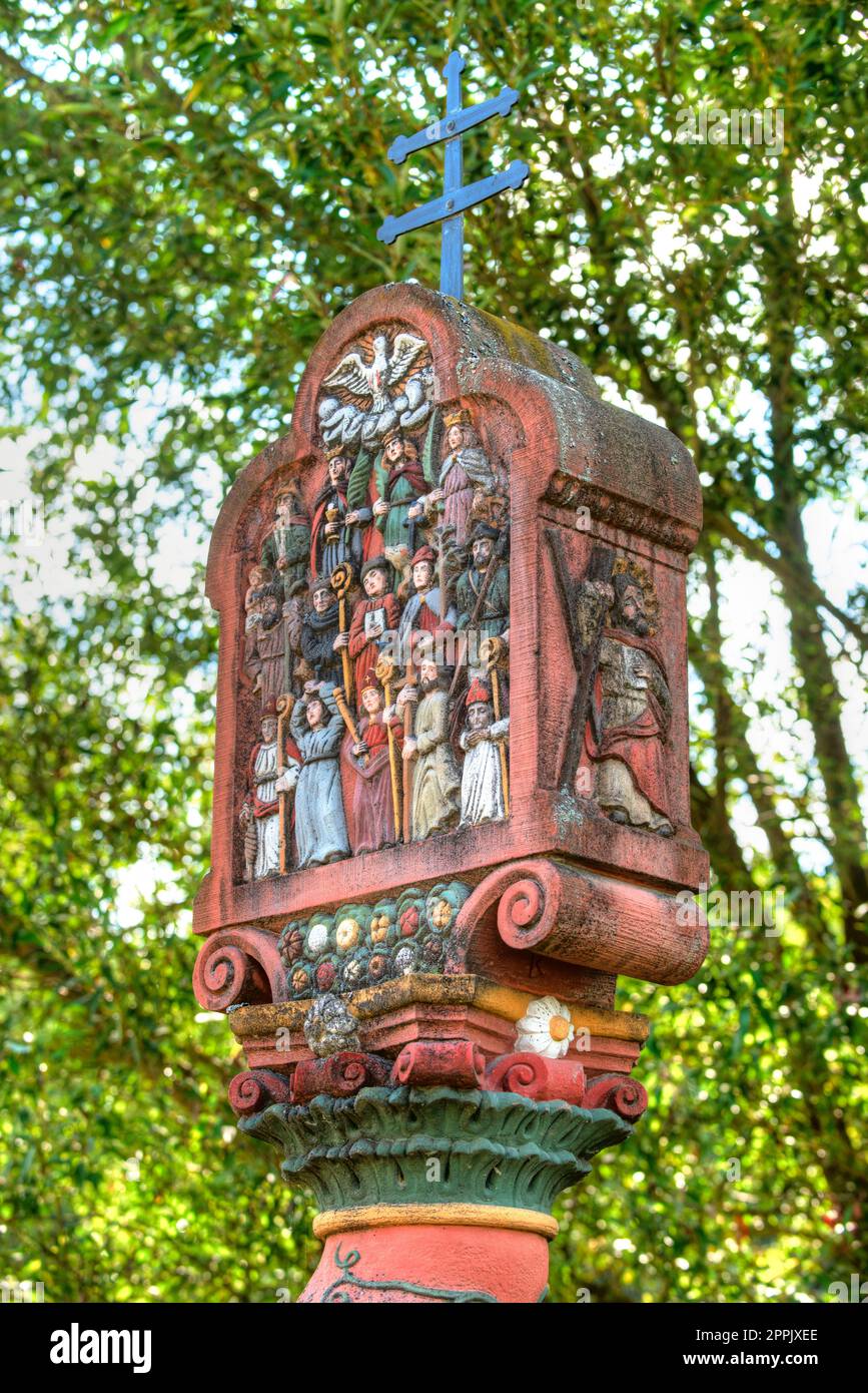 A wayside shrine colorfully decorated with Christian symbols and figures of saints in front of the green foliage of a tree Stock Photo