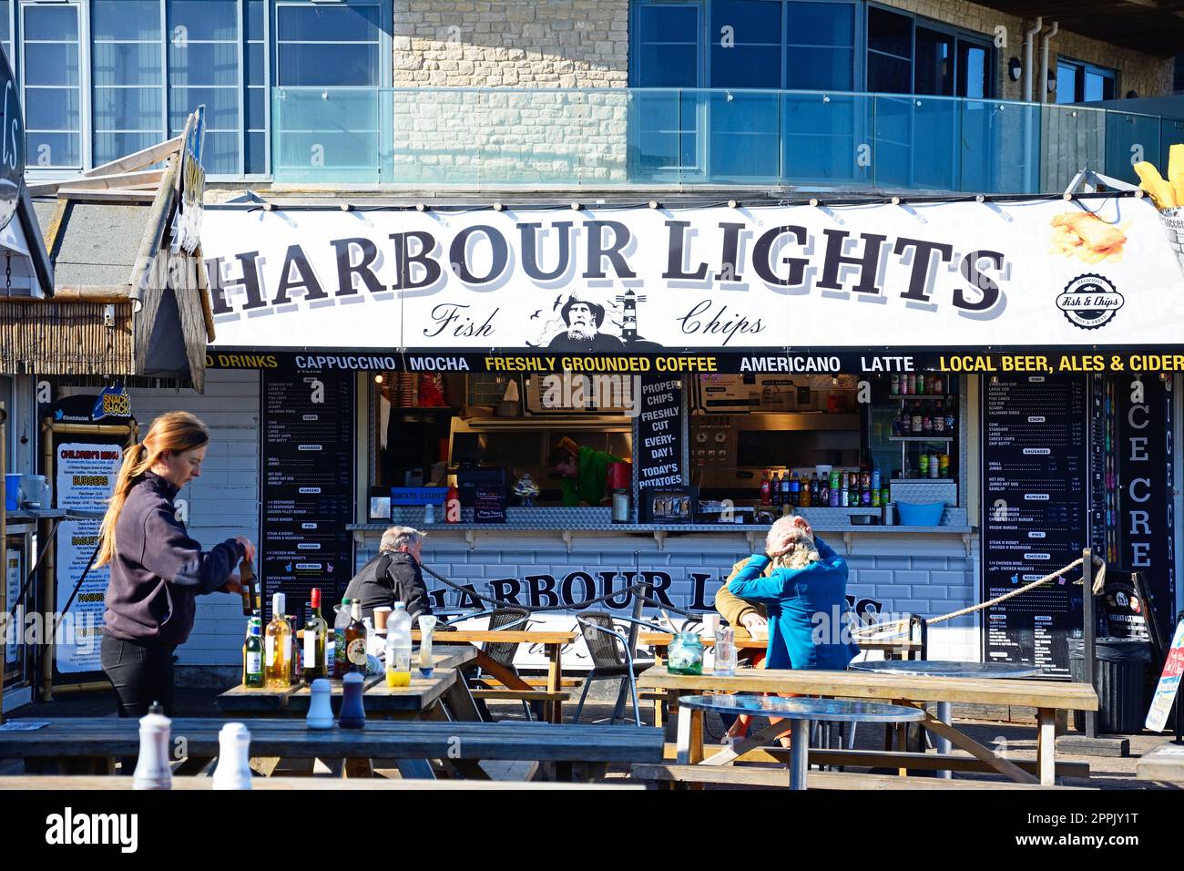 Tourists sitting outside the Harbour Lights cafe restaurant in the harbour area, West Bay, Dorset, UK, Europe. Stock Photo