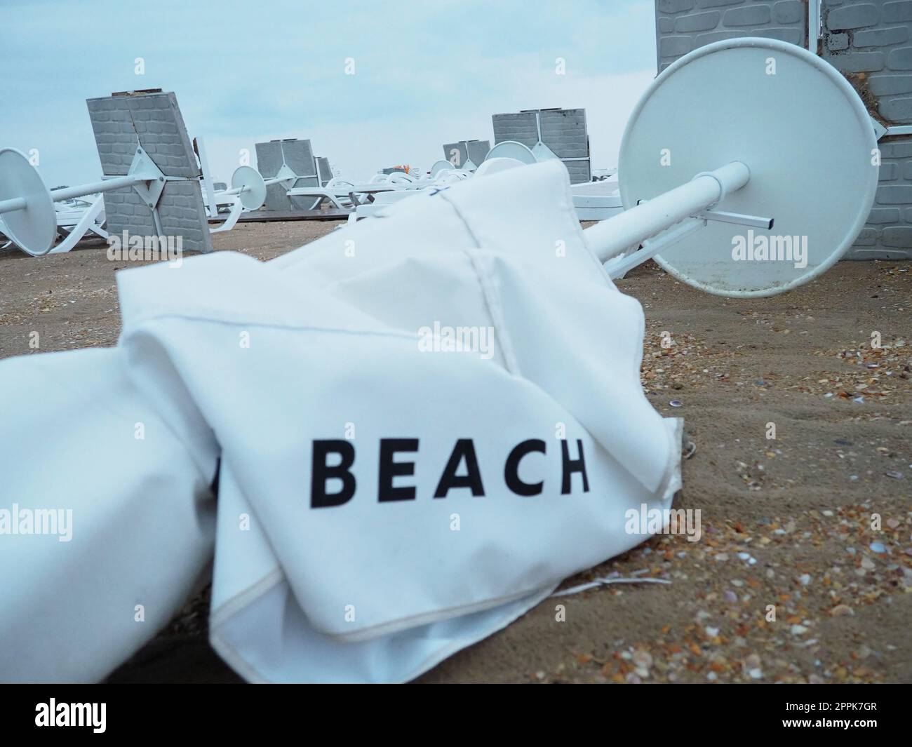 A beach umbrella lay folded on a sandy beach after bad weather. Storm at sea. End of the beach season due to typhoon and cyclone. Loungers, umbrellas and hotel furniture. Beach word written on white Stock Photo