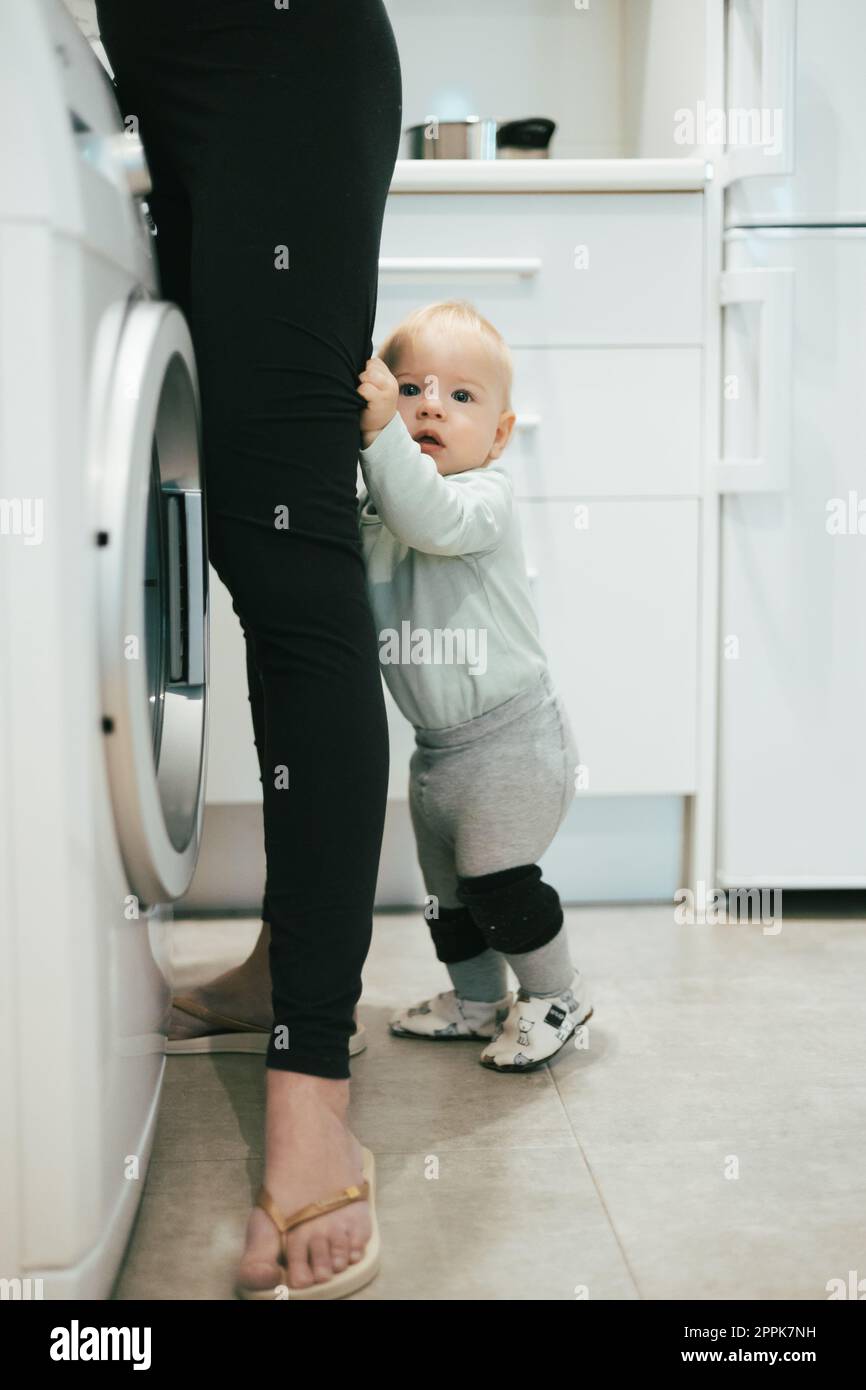 Little infant baby boy child hiding between mothers legs demanding her attention while she is multitasking, trying to do some household chores in kitchen at home. Mother on maternity leave. Stock Photo