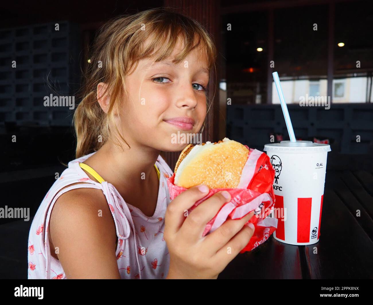 Anapa Russia 23 August 2021 A beautiful girl of 7 years old is eating at the KFC restaurant enjoying the food and smiling. Caucasian child holding a burger. Cola in a cardboard cup with lid and straw Stock Photo