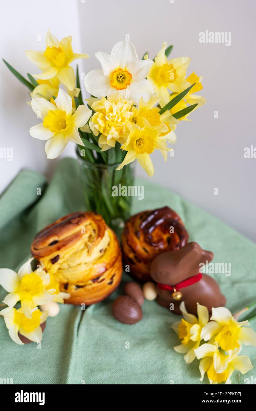 Homemade Easter traditional pastries lie on a green napkin along with daffodil flowers, rabbit, chocolate eggs. Easter baking and decor, vertical photo. Stock Photo