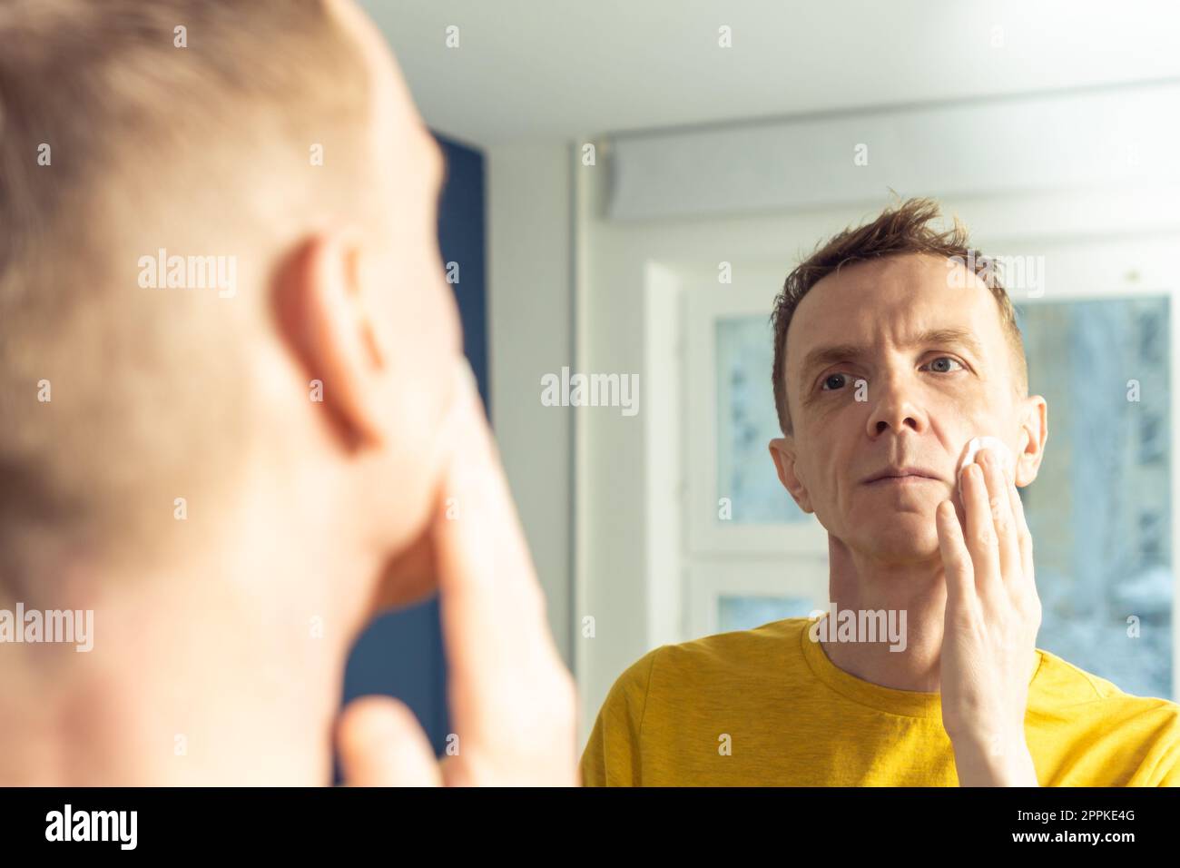 Mature man apply face tonic with cotton pad, then massage with hands. Male portrait in mirror. Face cleansing, washing. Stock Photo