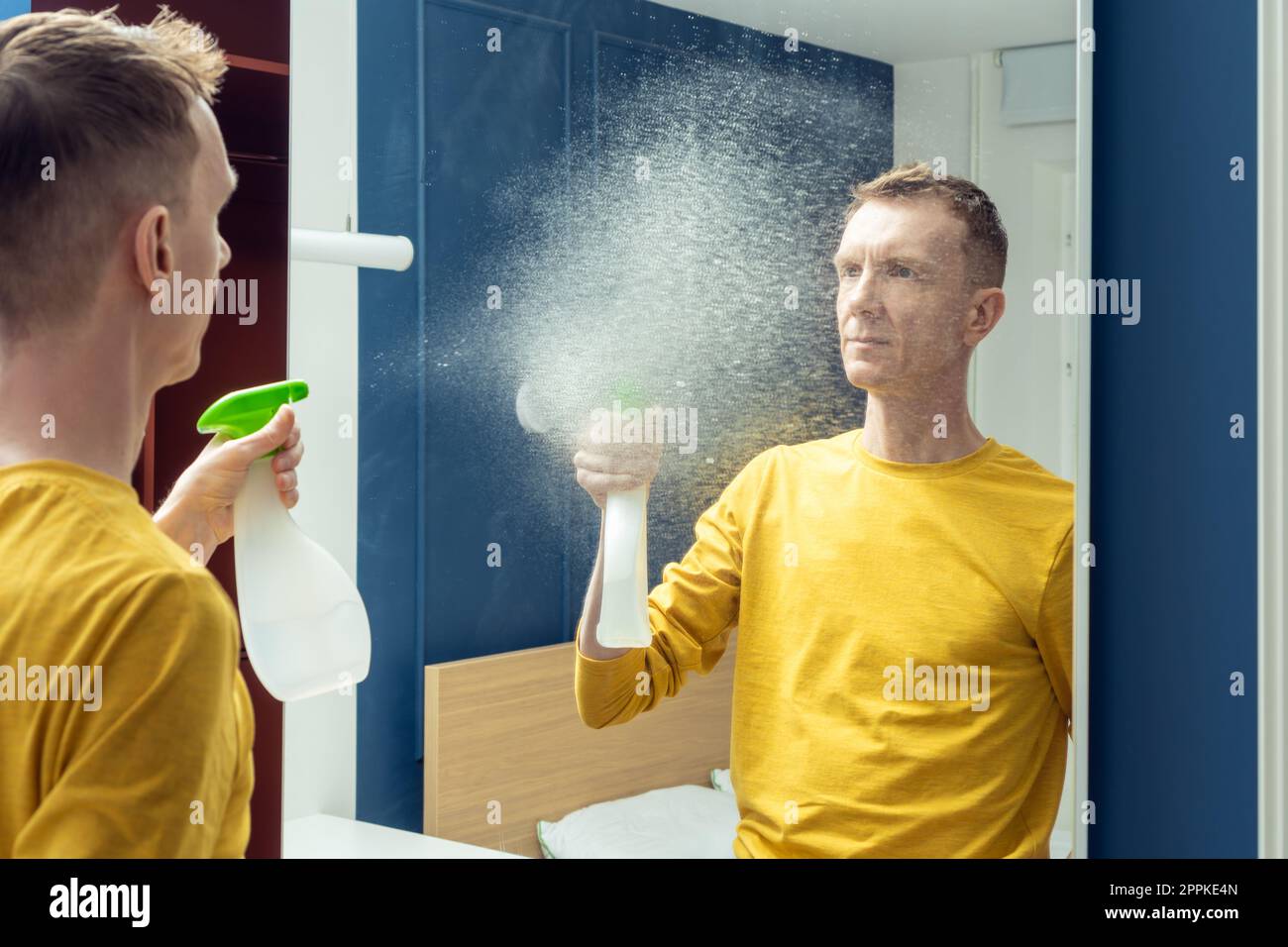 Adult man hold bottle of cleanser and spray splashes on mirror of sliding wardrobe door. Wash mirror with cleaners. Stock Photo