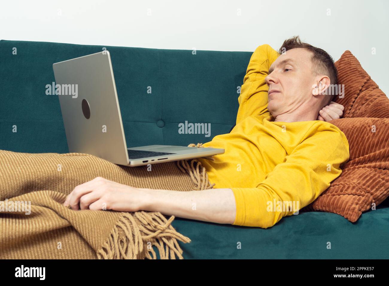 Tired man lie on sofa and watch movie on laptop in living room. Adult person reclining on pillow under blanket. Stock Photo