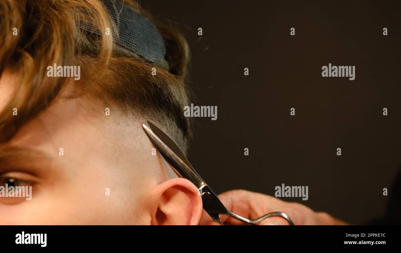 Close up Barber master hairdresser does hairstyle and style with scissors and comb. Concept Barbershop. side view in dark key lightning with warm light Stock Photo