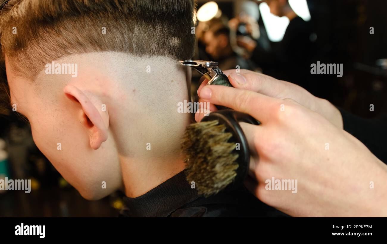 Shot of a handsome barber giving a haircut to his client using trimmer. Hairdresser service in a modern barbershop in a dark key lightning with warm light side view Stock Photo