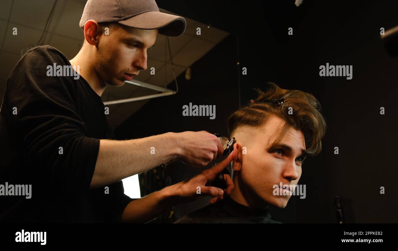 men's hairstyling and haircutting with hair clipper in a barber shop or hair salon. Hairdresser service in a modern barbershop in a dark key lightning with warm light side view Stock Photo