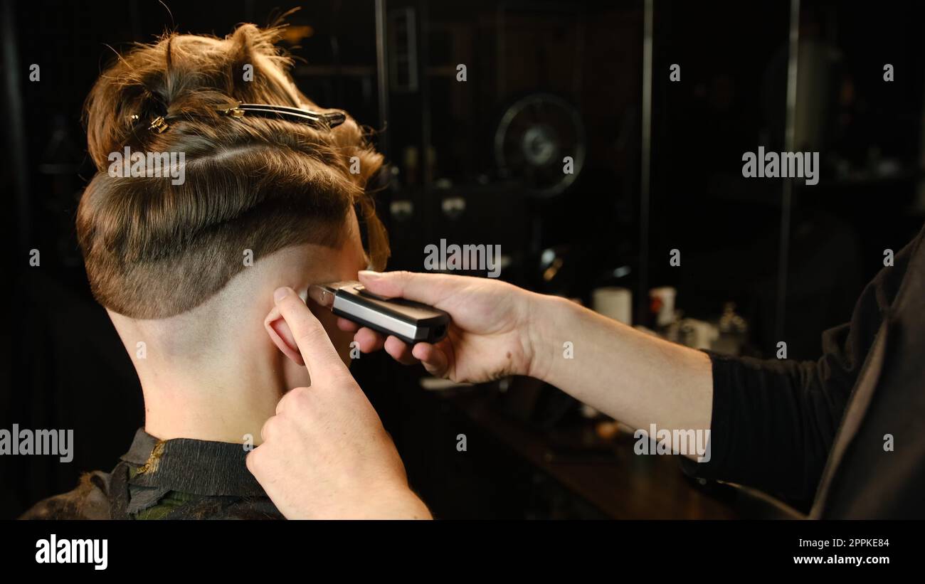 men's hairstyling and haircutting with barber shaving machine, hair cut for male. Hairdresser service with shaver in a modern barbershop in a dark key lightning with warm light back view Stock Photo