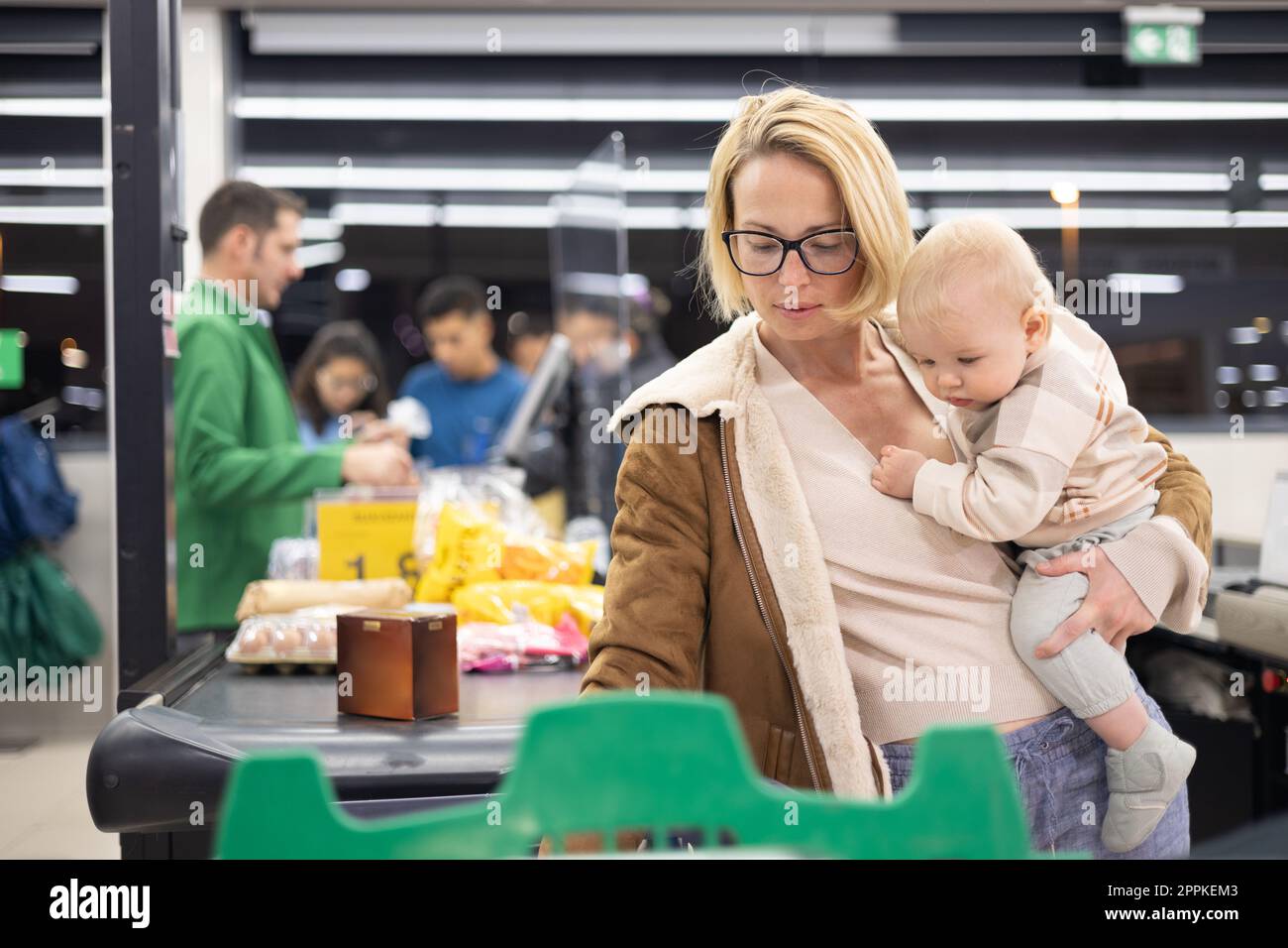 Mother shopping with her infant baby boy, holding the child while stacking products at the cash register in supermarket grocery store. Stock Photo