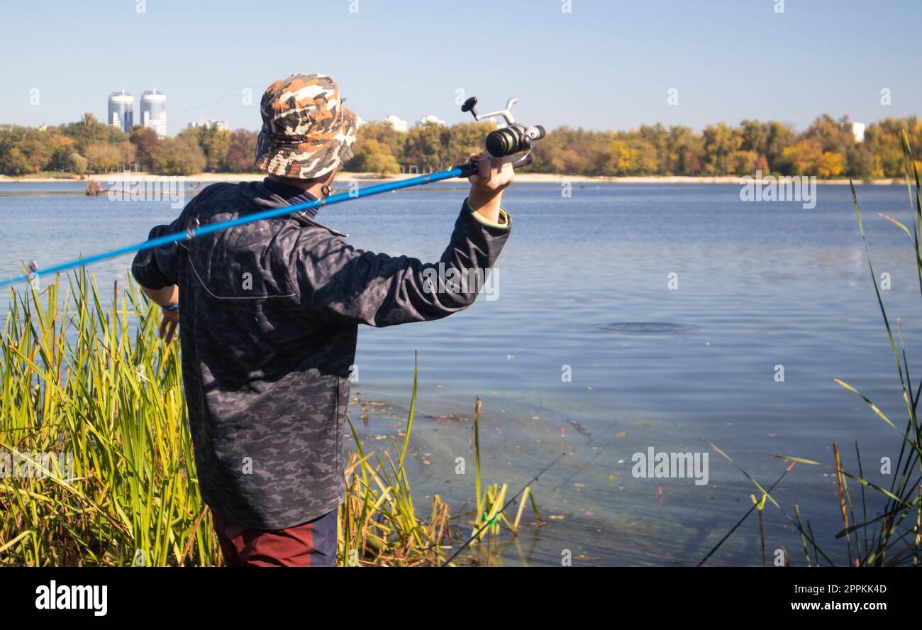 A male fisherman threw a spinning rod into a lake or river on a sunny day. Shore fishing. A fisherman on the river bank holds a spinning rod in his hand. The concept of country rest. Fishing article. Stock Photo