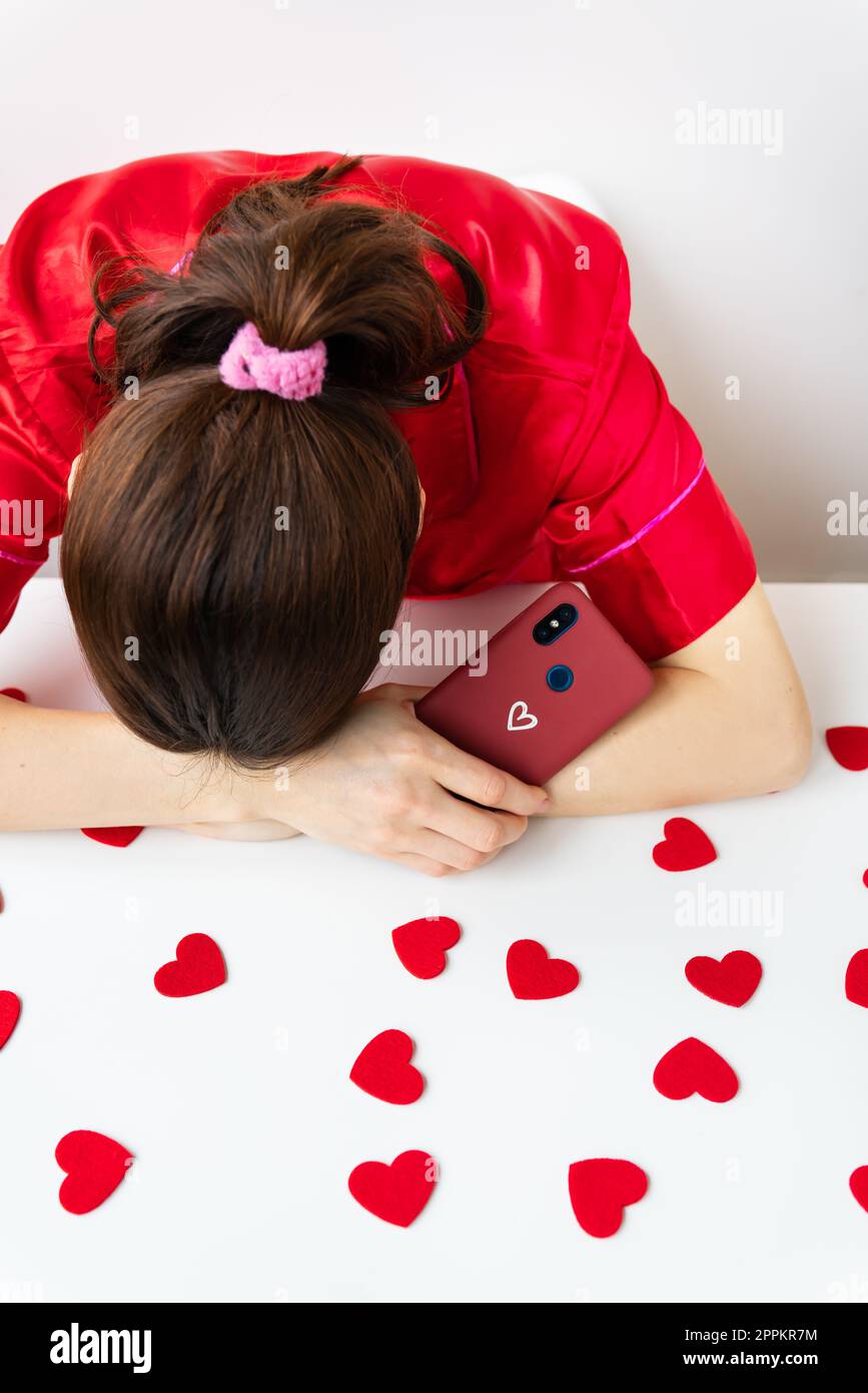 The girl is sad, puts her head in her hands, holds a smartphone in a cherry case with a heart, waiting for a text message or an invitation to a date. Valentine's day and dating concept. Stock Photo