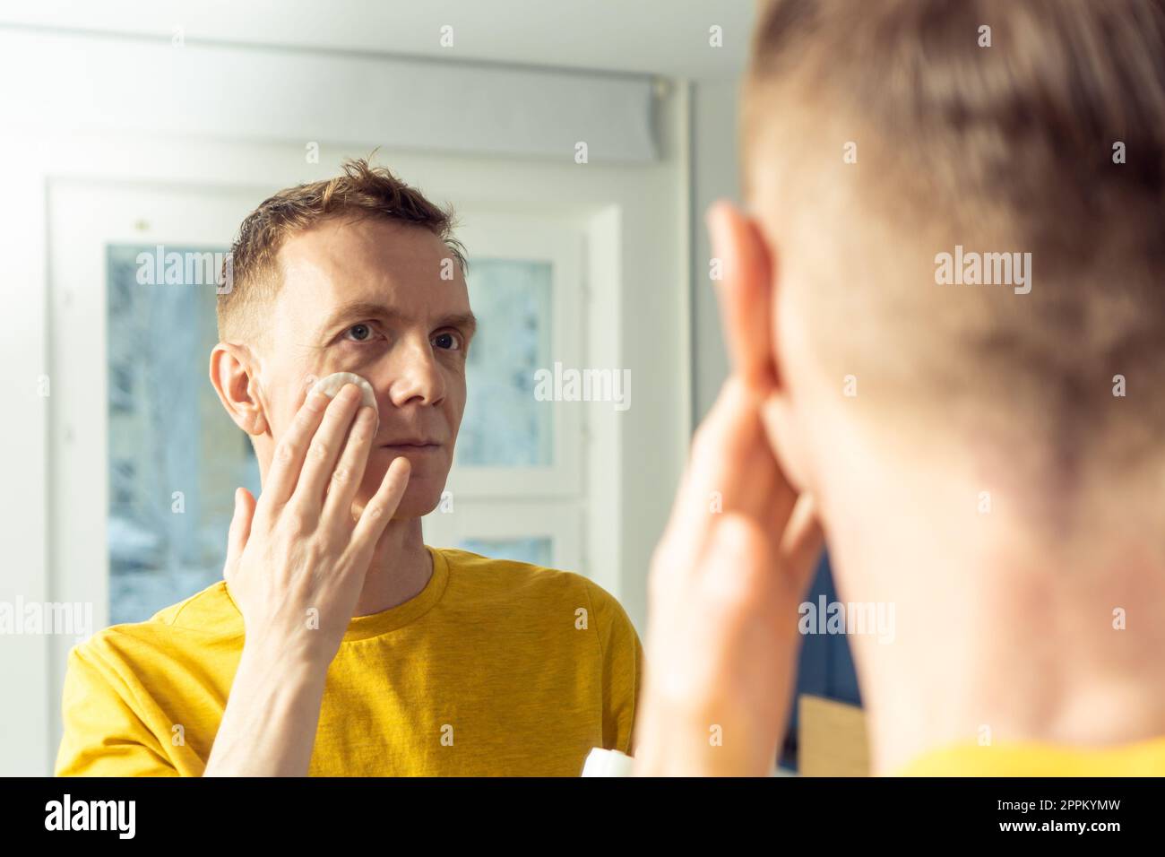 Middle aged man apply face tonic with cotton pad, then massage with hands. Male portrait in mirror. Face cleansing. Stock Photo