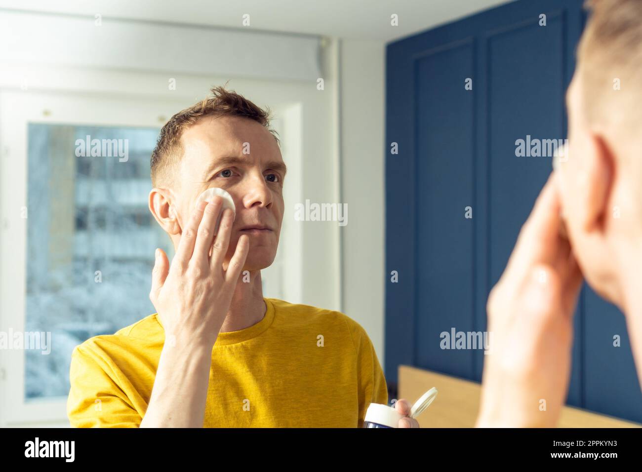 Mature man apply face tonic with cotton pad, then massage with hands. Male portrait in room mirror. Face cleansing. Stock Photo