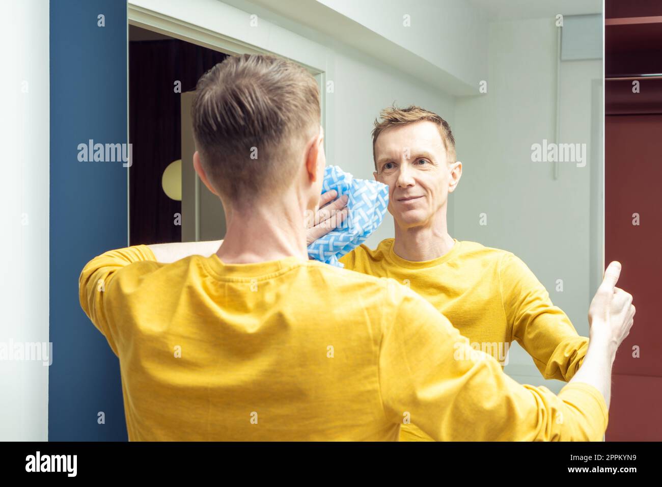 Happy adult man wiping mirror of sliding wardrobe door with dry rag, back view. Polish mirror with household cloth. Stock Photo