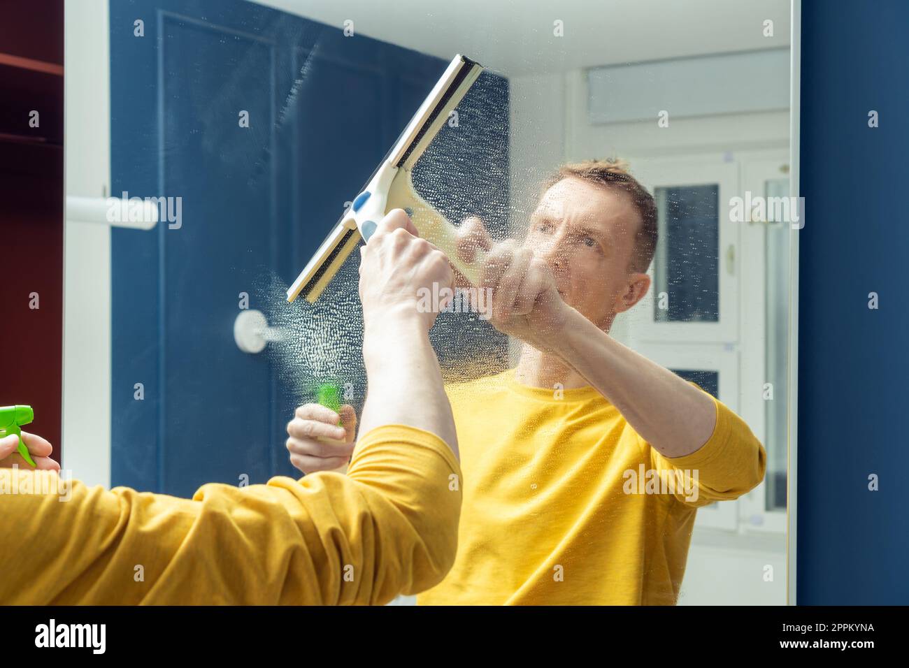 Middle aged man spray cleanser on mirror of sliding wardrobe door and wipe with plastic brush. Polish mirror. Stock Photo