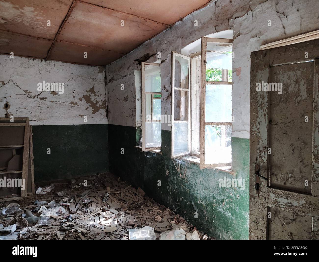 Inside an abandoned house. Scattered trash on the floor. Green paint, cracks and cobwebs on the wall. The white upper half of the wall. Open windows through which sunlight shines through. Old door Stock Photo