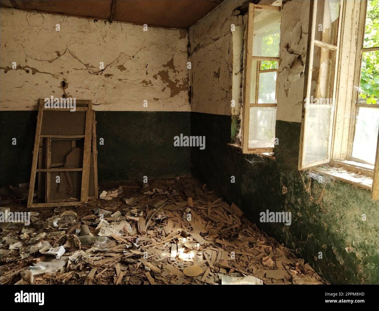 Inside abandoned house. Scattered trash on the floor. Green paint, cracks and cobwebs on the wall. The white upper half of the wall. Open windows through which sunlight shines through Frames removed Stock Photo