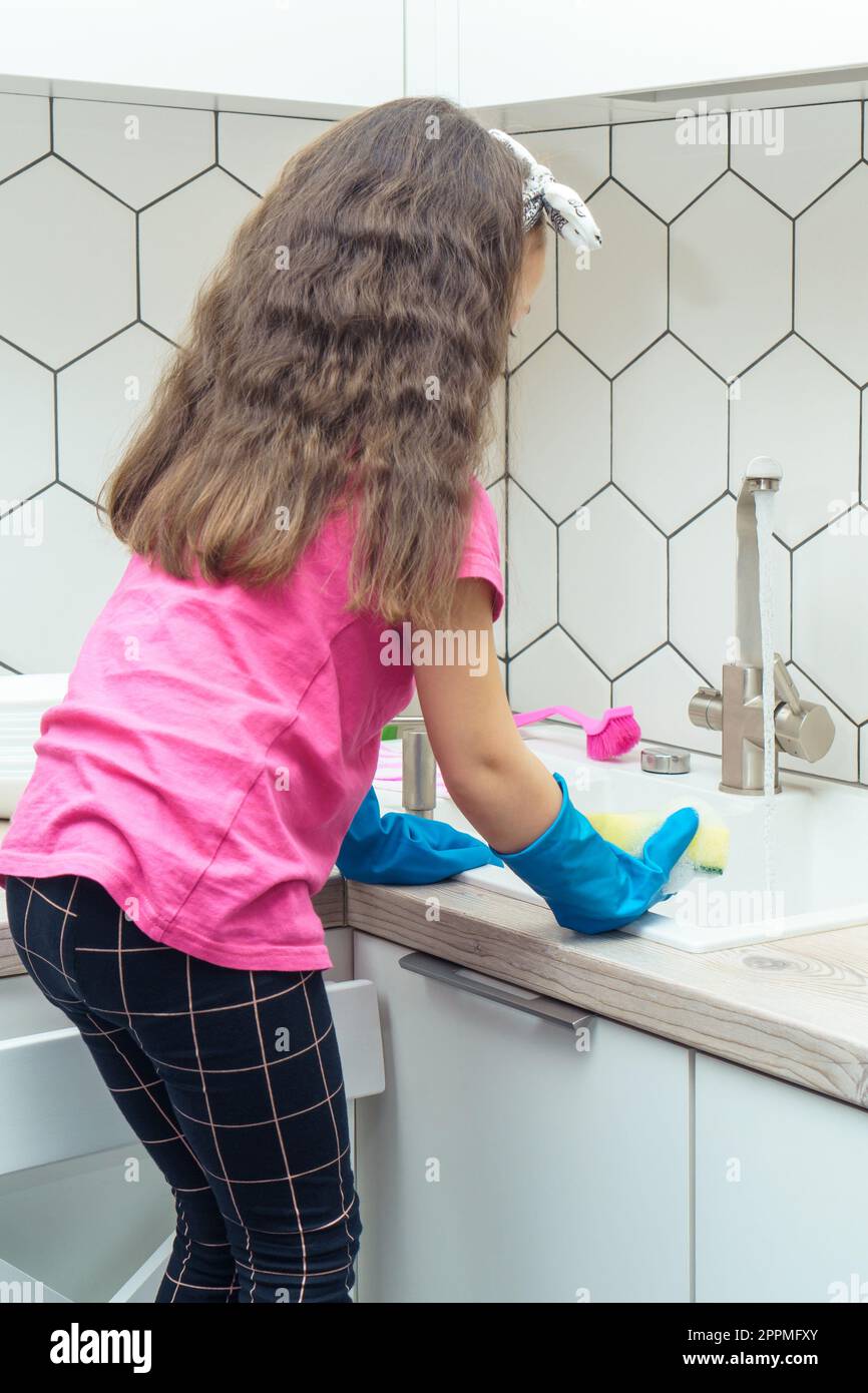Little girl in protective gloves wash up dishes with sponge in kitchen sink standing on chair. Tidying up kitchen. Stock Photo