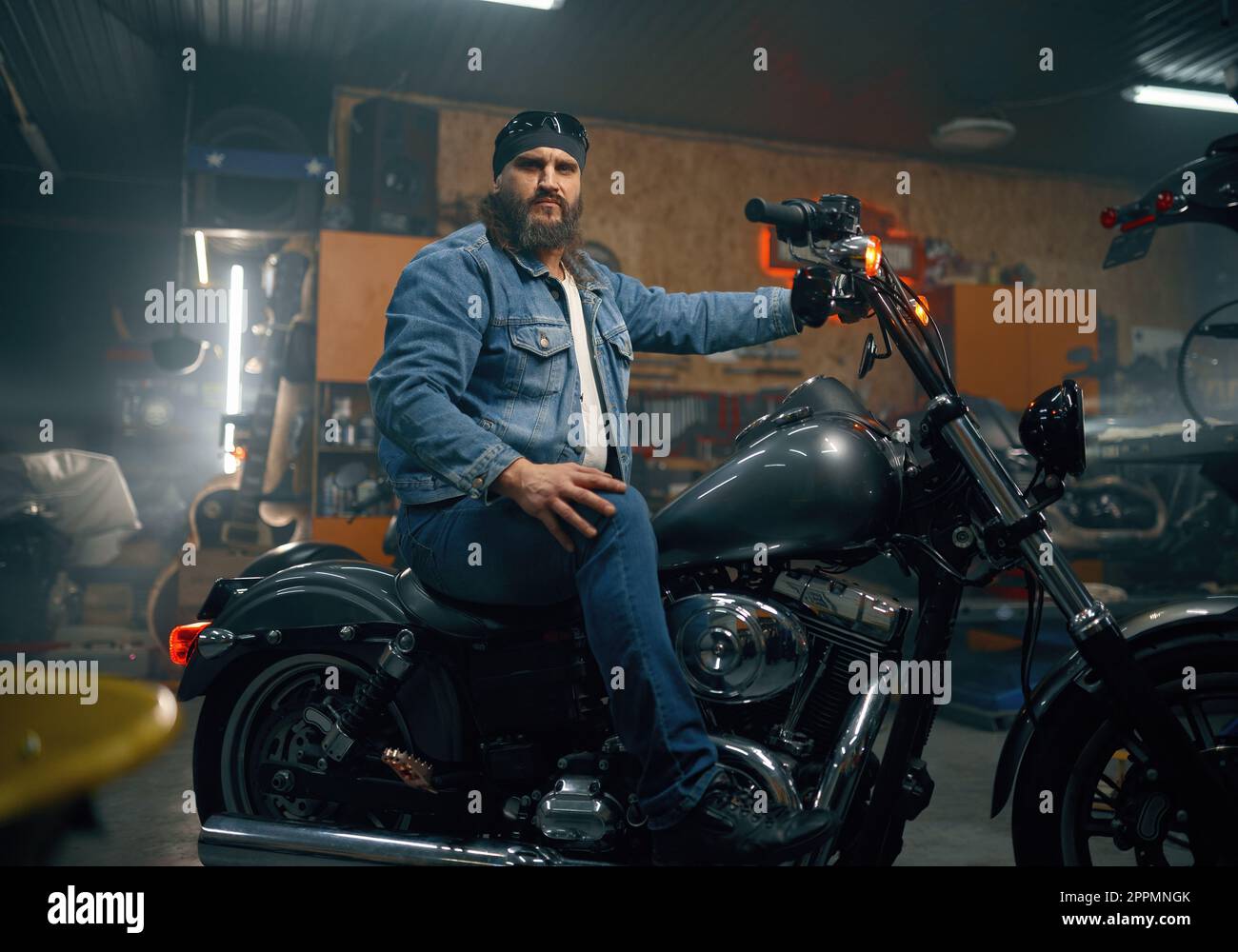 Portrait of mature bearded biker on motorcycle over garage interior Stock Photo