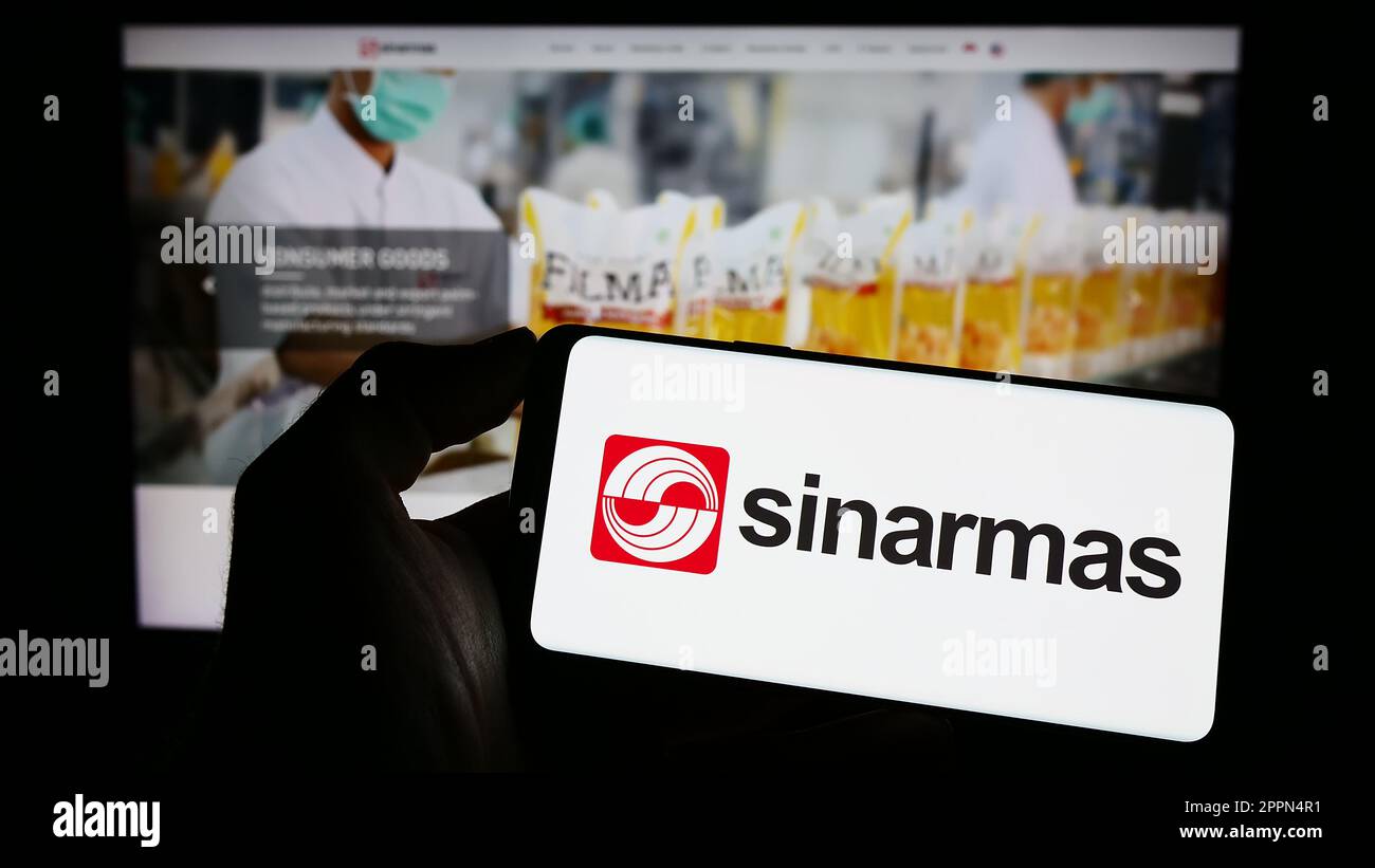 Person holding mobile phone with logo of Indonesian conglomerate Sinar Mas Group on screen in front of business web page. Focus on phone display. Stock Photo