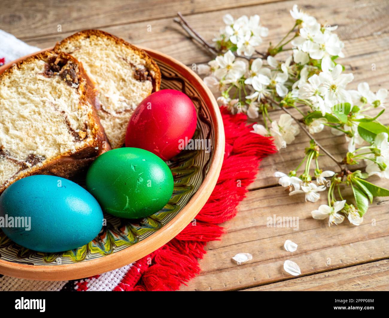 red Easter eggs next to flowering branches and plate with sweet sponge cake or cozonac on wooden table Stock Photo