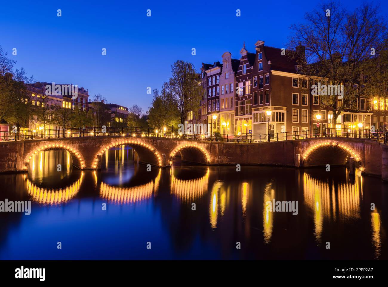 A tranquil evening in Amsterdam, illuminated by the soft glow of dusk. A beautiful arch bridge reflects on a canal amidst stunning architecture and ri Stock Photo
