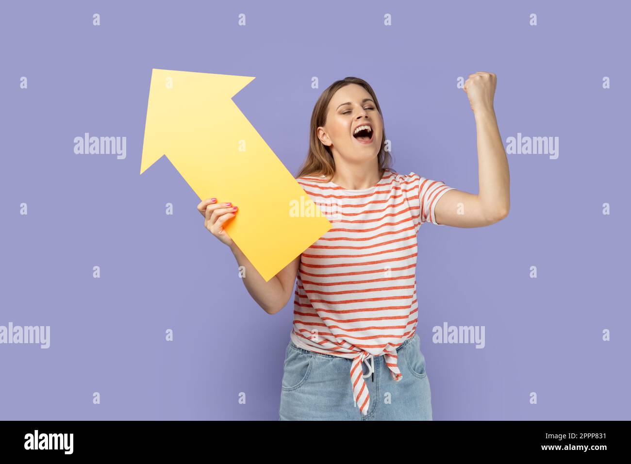 Portrait of extremely happy woman wearing striped T-shirt holding big yellow arrow pointing aside and clenched fist, celebrating victory, increase. Indoor studio shot isolated on purple background. Stock Photo