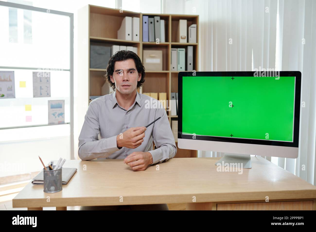 Young male business coach pointing at green screen of computer while sitting by workplace and making presentation to audience Stock Photo