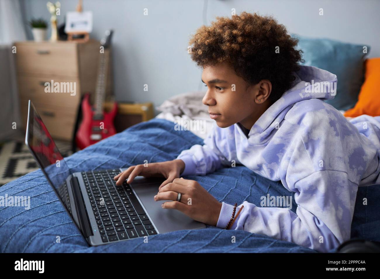 Cute teenage boy in hoodie relaxing on bed with laptop while watching curious online movie or communicating in video chat at leisure Stock Photo