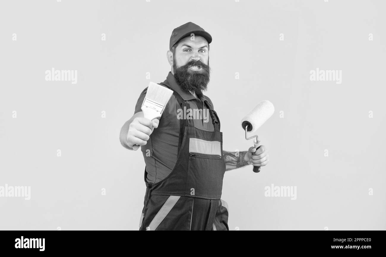 aggressive man in work clothes hold paint roller and brush on yellow
