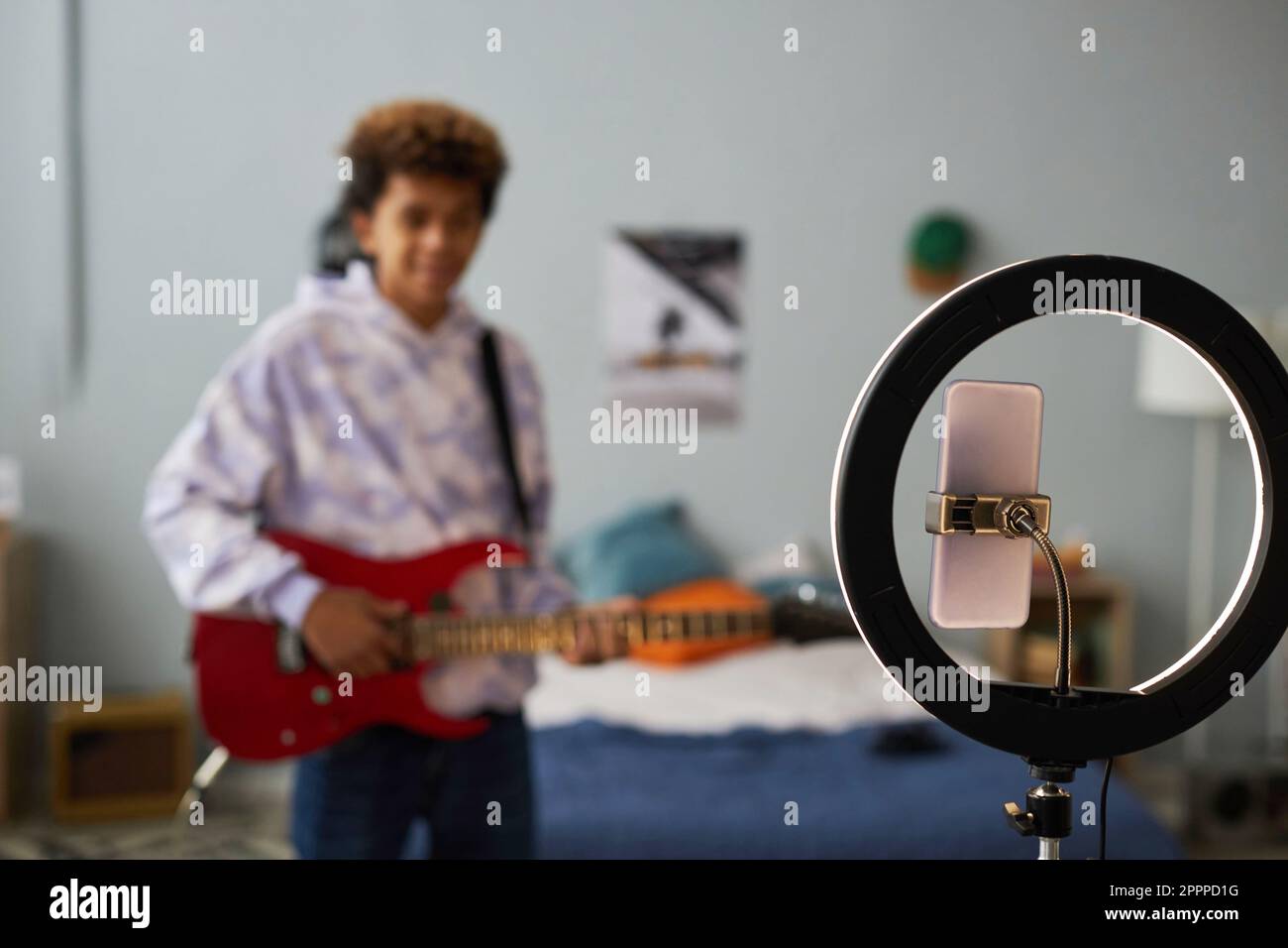 Focus on round frame with smartphone on holder during video recording against teenage guy playing electric guitar in his bedroom Stock Photo