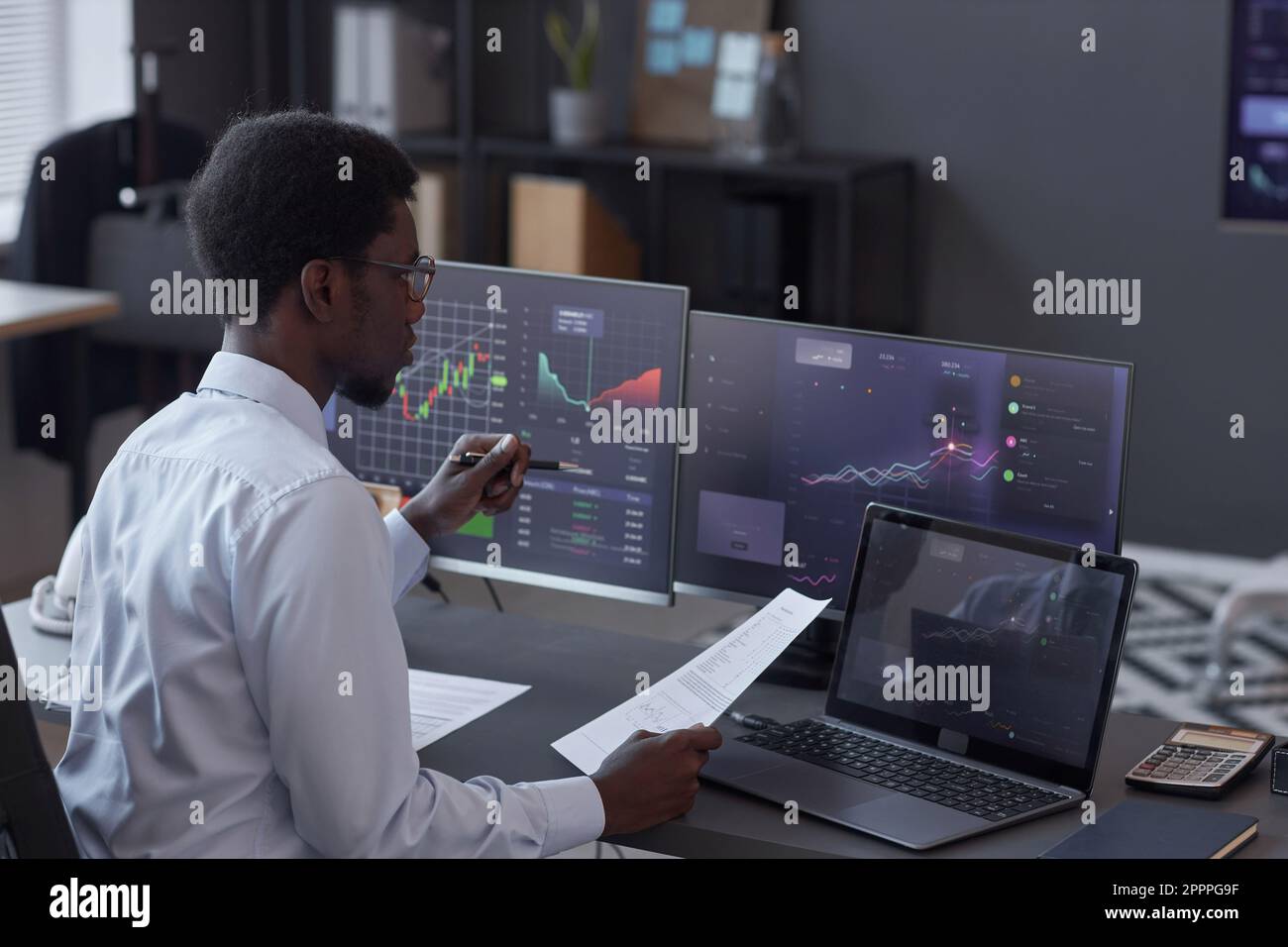 African American broker analyzing financial market while sitting at table in front of monitors with charts in office Stock Photo