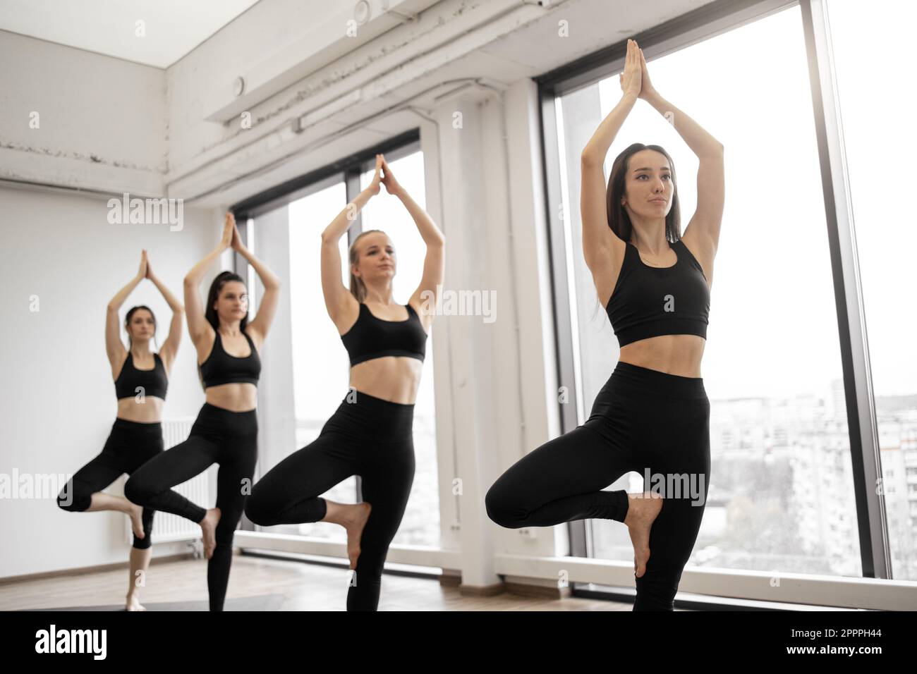 Group of athletic females in tight activewear standing in Tree Pose in white room of sports club. Young yoginis strengthening posture with help of Vrikshasana exercise during training session indoors. Stock Photo