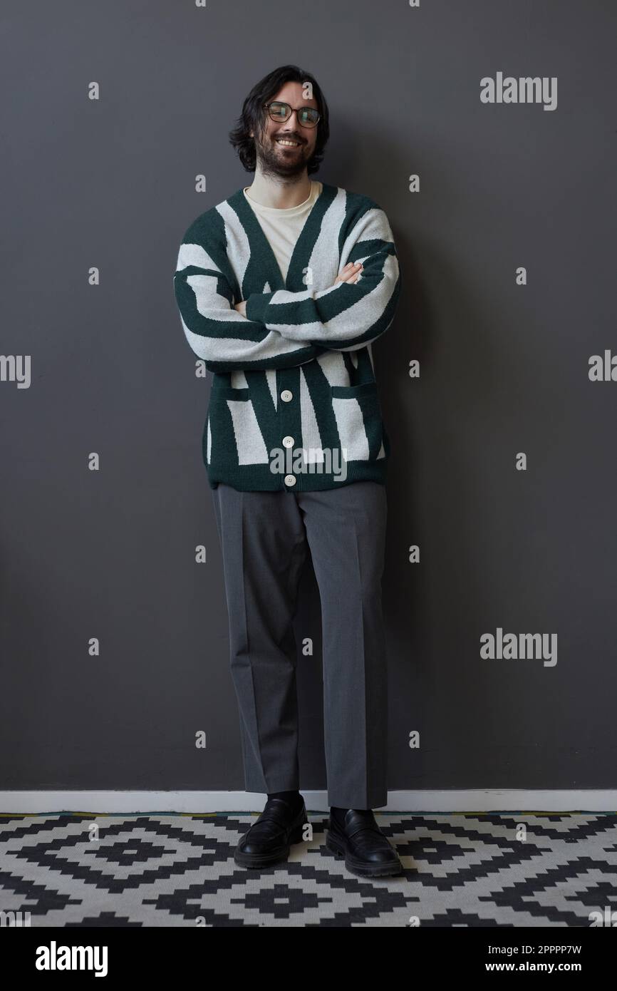 Vertical image of stylish young man in cardigan smiling at camera standing against grey wall Stock Photo