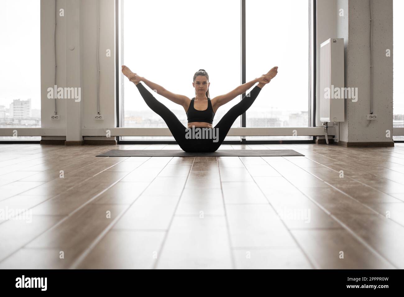 Front view of beautiful fitness model in black sportswear doing stretching while sitting on floor of white capacious studio. Caucasian dark haired lady balancing on yoga mat during everyday training. Stock Photo